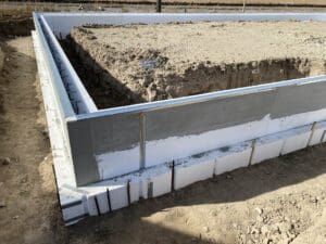The image shows the foundation of a building under construction, with a square-shaped concrete base surrounded by insulated concrete forms. The area within the foundation is filled with compacted soil, ready for further construction.