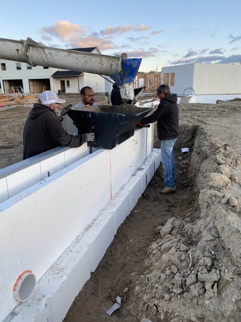 Three workers pour concrete into a mold with a large hose on a construction site. They are surrounded by insulated concrete forms and soil. Buildings are visible in the background under a cloudy sky.