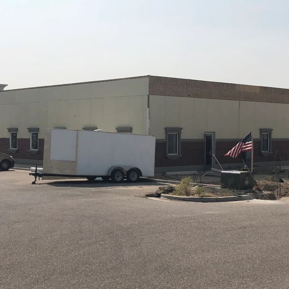 A large, unfinished building in a parking lot with a construction trailer parked in front. The structure lacks a completed facade, and the American flag is mounted nearby. The sky is overcast.