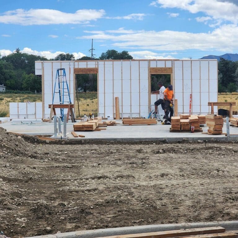 Construction site with workers assembling a building frame. The structure has white panels and wooden beams. A blue ladder is set up in front. The area is surrounded by dirt and open fields, with trees and mountains in the background under a partly cloudy sky.