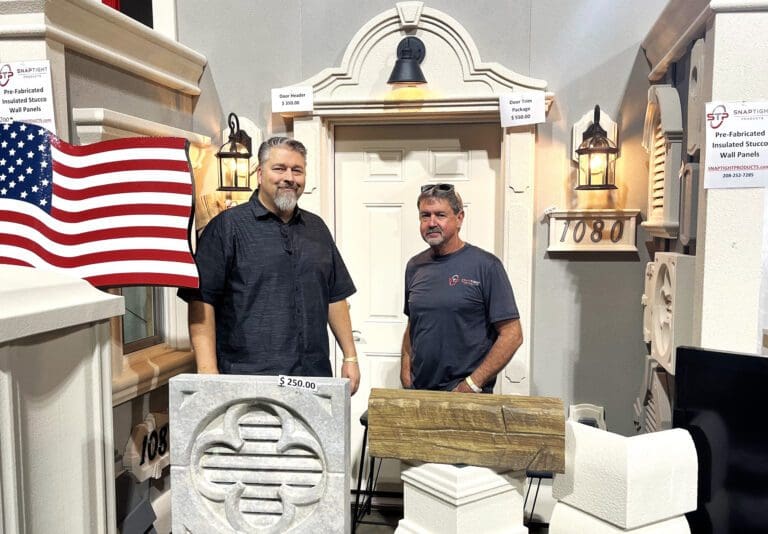 Two men standing in front of a display for pre-fabricated insulated siding panels. They are surrounded by decorative insulated building products and an American flag. Both are smiling, with price tags visible on some items.