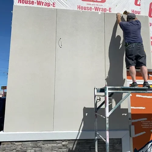A person on scaffolding applies plaster to a wall beneath Grip-Rite house wrap, with the lower section covered in Prefabricated Wall Panels, outdoors on a sunny day.